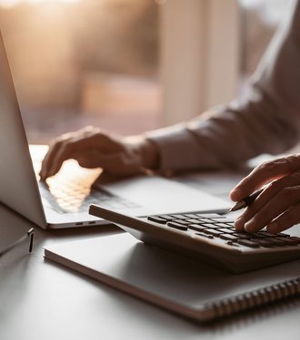 Mensch arbeitet am Tisch mit Laptop und Taschenrechner